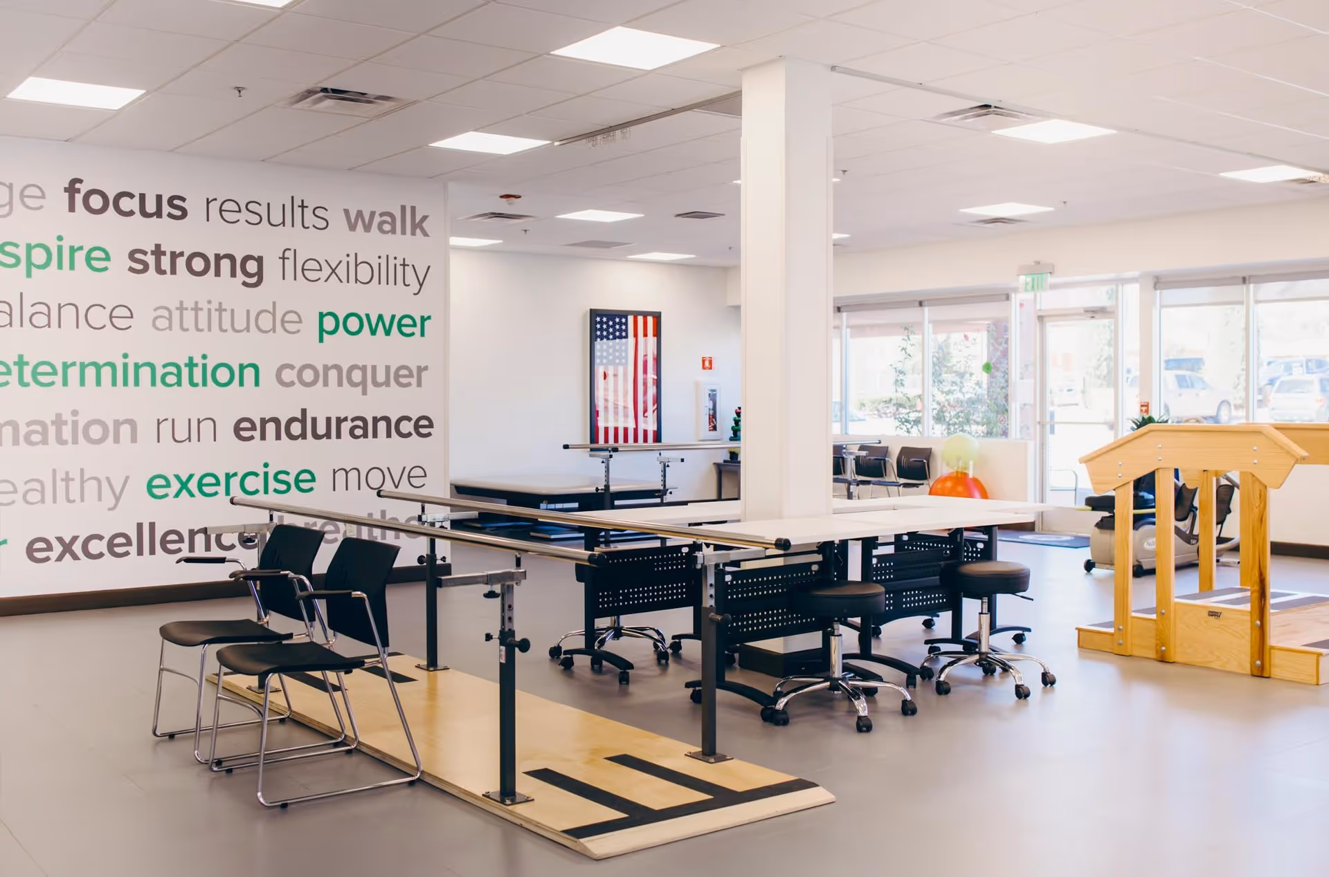 A bright rehabilitation or physical therapy room with parallel bars for walking exercises, chairs, stools, and a wooden ramp. The walls have motivational words like focus, inspire, strong, power, determination, endurance, and exercise. Large windows allow natural light to fill the space.