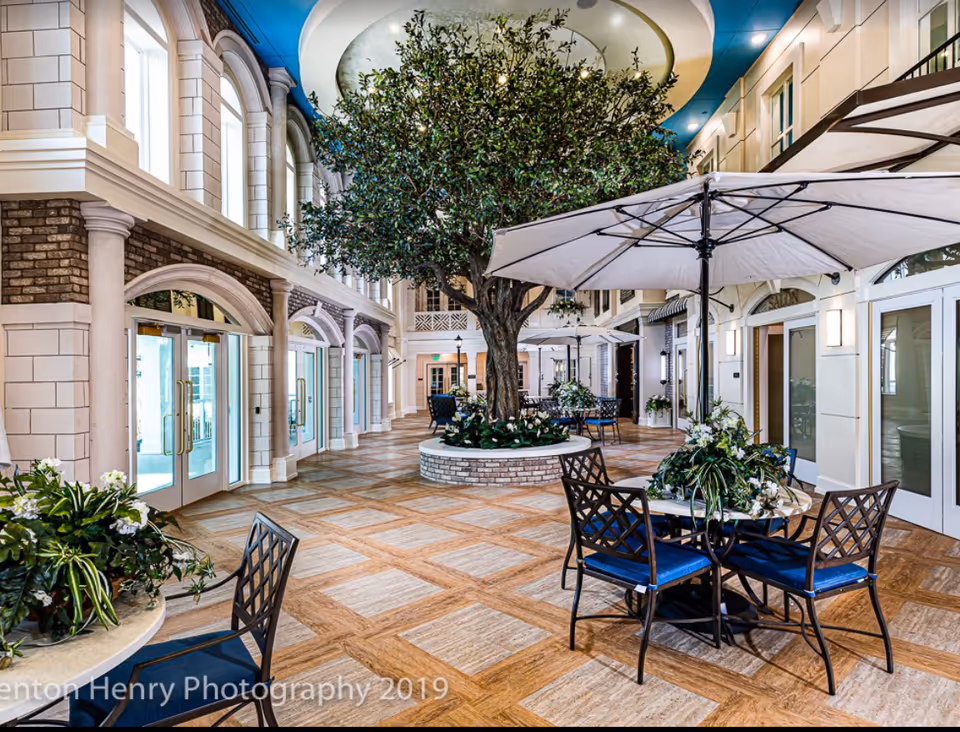 Indoor common area with a large artificial tree in the center surrounded by tables with umbrellas and chairs. The space features high ceilings with large windows, decorative columns, and a tiled floor with a wood pattern. There are plants on the tables and a bright, airy atmosphere.