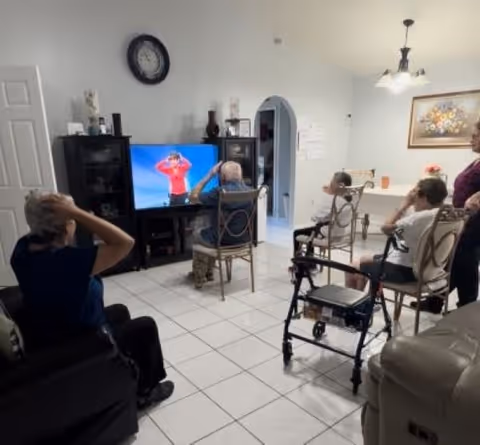 Several elderly residents sit in a living room following an exercise program playing on a television.