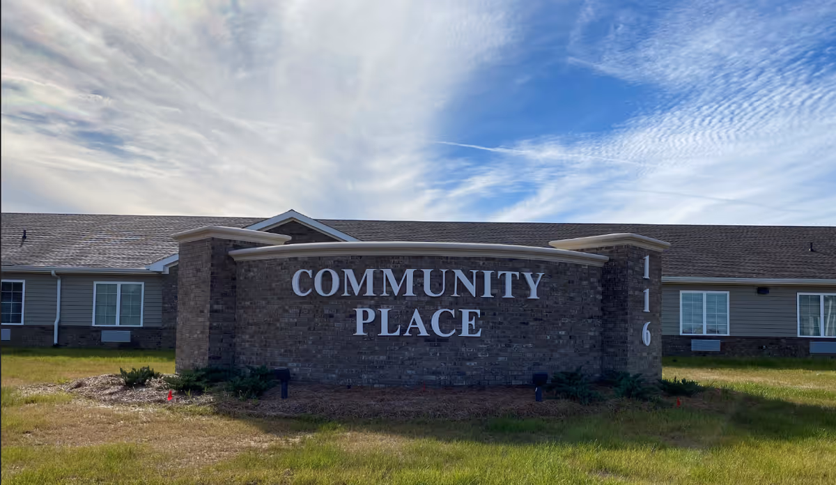 A large brick sign in front of a single-story building with the words COMMUNITY PLACE in white letters. The building has a gray roof and beige siding with several windows. The sky above is partly cloudy with blue patches.