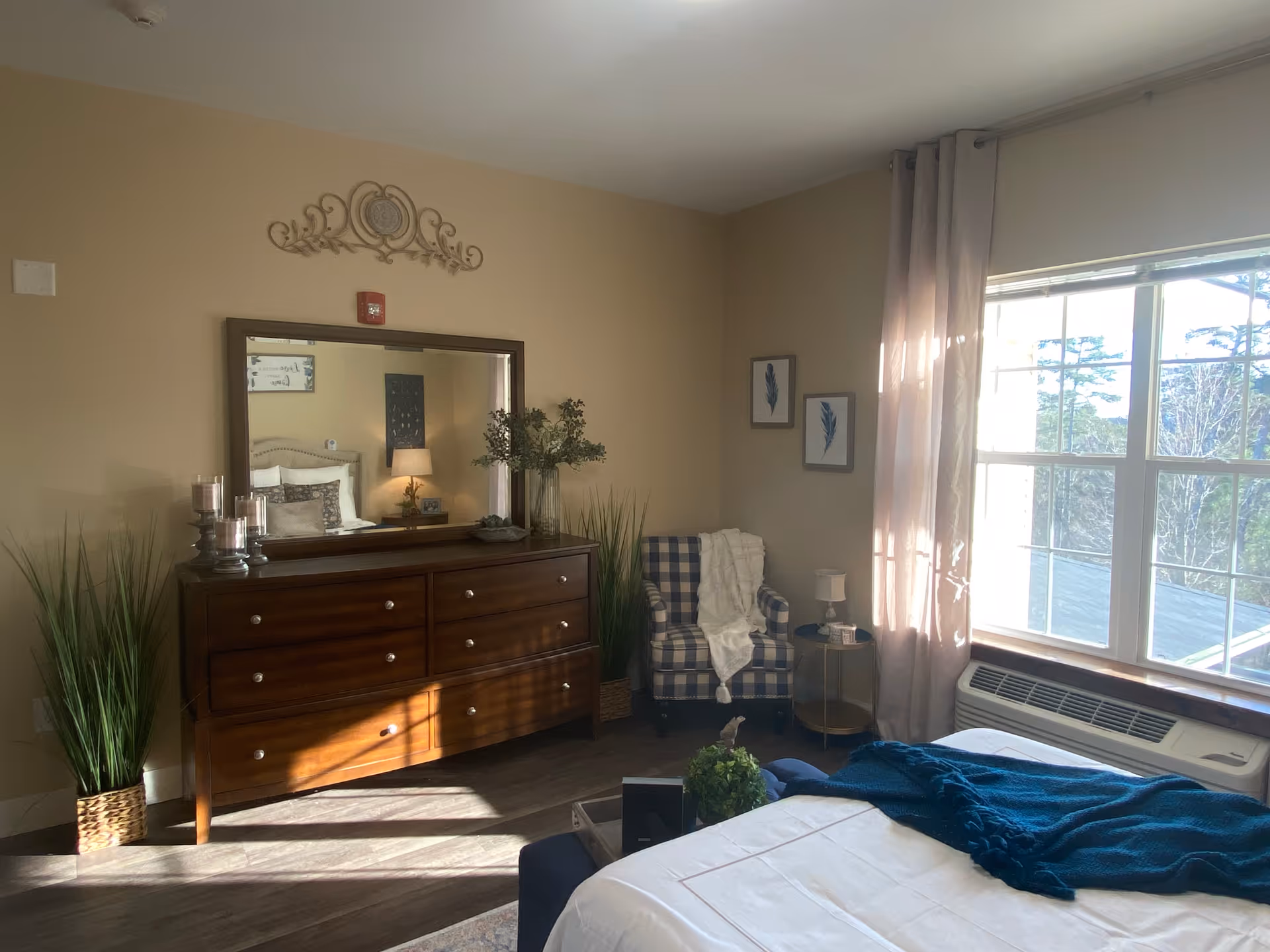A cozy bedroom with a large window letting in natural light. The room features a wooden dresser with a large mirror above it, two tall green plants on either side, a checkered armchair with a white throw blanket, and a small round side table with a lamp. The bed is partially visible with white bedding and a dark blue throw blanket.