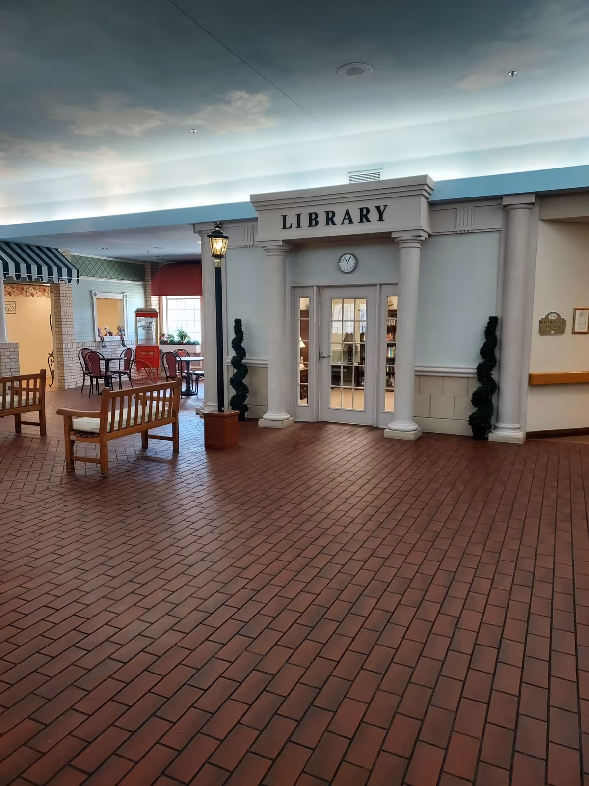 Indoor view of a senior living facility lobby with a mock 'LIBRARY' entrance, benches, a lamp post and brick-tile floor.