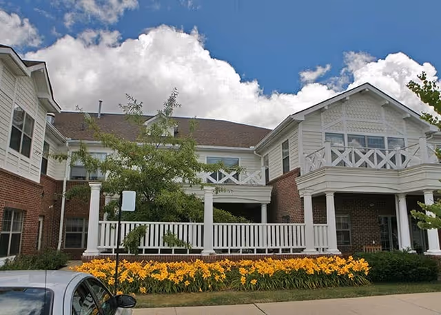 Front exterior of a two-story brick and white-siding building with a wraparound porch, balcony, and a bed of yellow flowers in front.