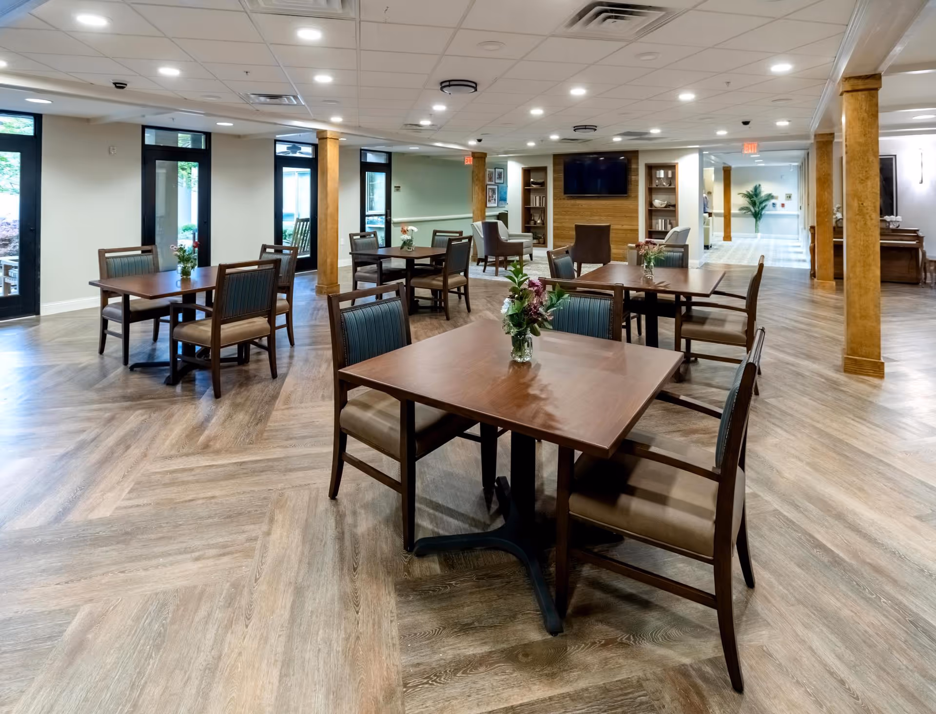 Spacious senior living dining area with several wooden tables and chairs, floral centerpieces, and a seating area with a TV.
