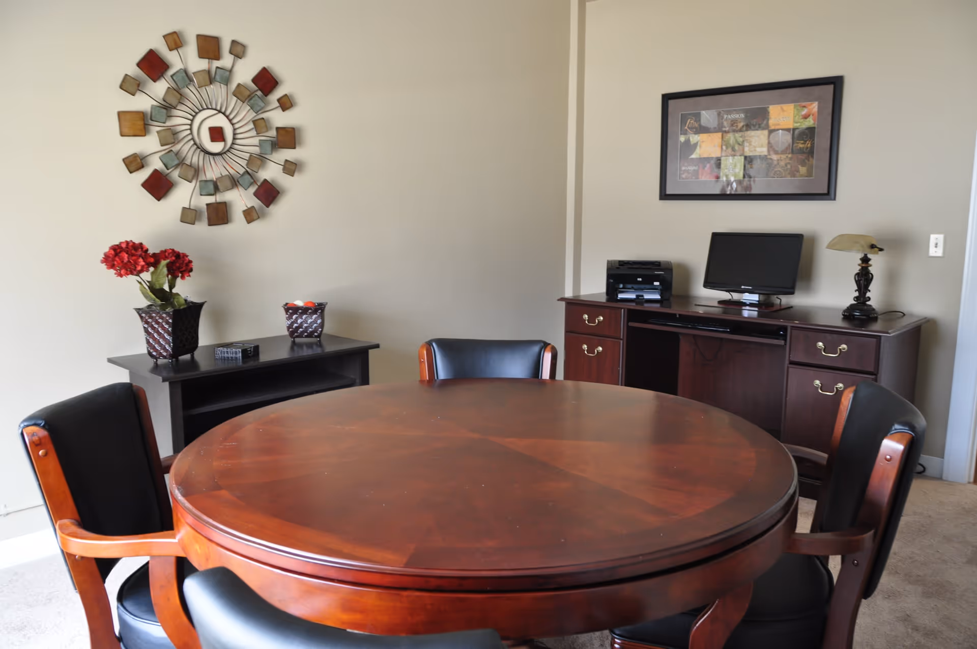A round wooden table surrounded by four black cushioned chairs in a room with beige walls. Against the wall is a dark wooden desk with a computer monitor, printer, and a decorative lamp. Above the desk hangs a framed artwork. On the left side, there is a small table with two decorative pots, one with red flowers. A decorative wall piece with square elements is mounted on the wall above the small table.