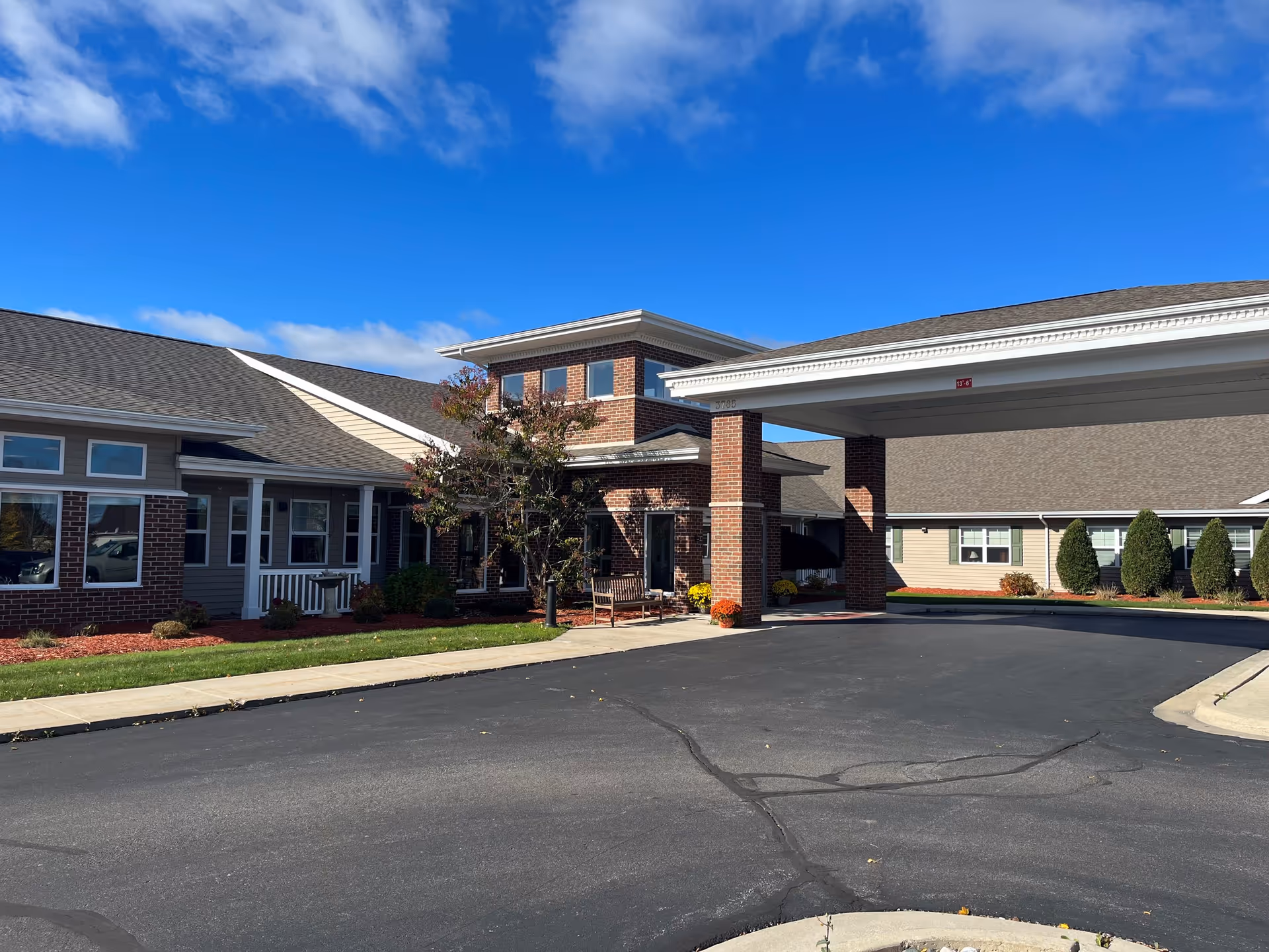 Exterior view of Traditions of Saginaw Main Campus building entrance with a covered drop-off area, brick and siding facade, landscaped bushes and trees, and a clear blue sky.