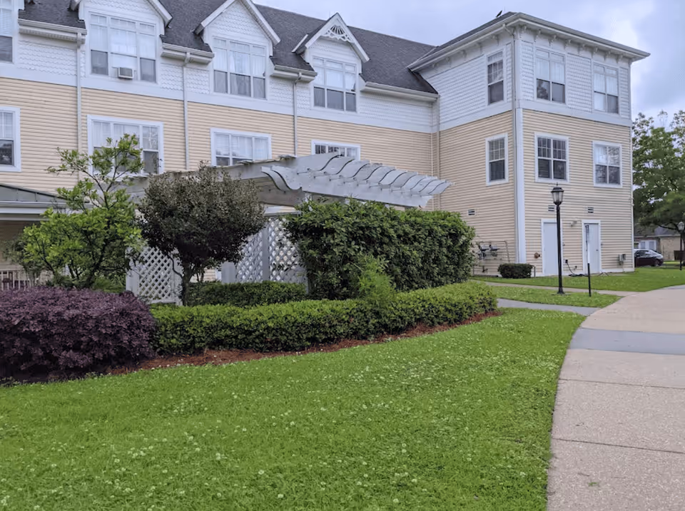 Exterior view of a multi-story residential building with beige and white siding. In front of the building, there is a well-maintained lawn with green grass, various shrubs, and a white wooden pergola. A paved walkway curves along the right side of the image, and a black lamppost is visible near the building.