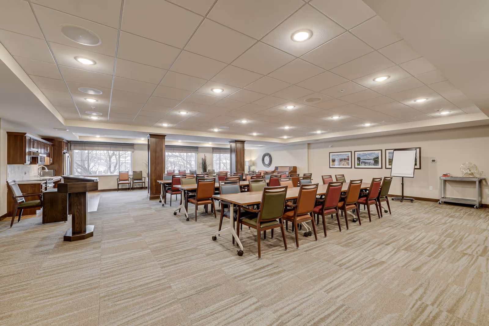A spacious, well-lit meeting or dining room with multiple tables and chairs arranged in rows. The room features a carpeted floor, a podium, a kitchen area with wooden cabinets and stainless steel appliances, large windows with blinds, framed pictures on the wall, and a flip chart stand.