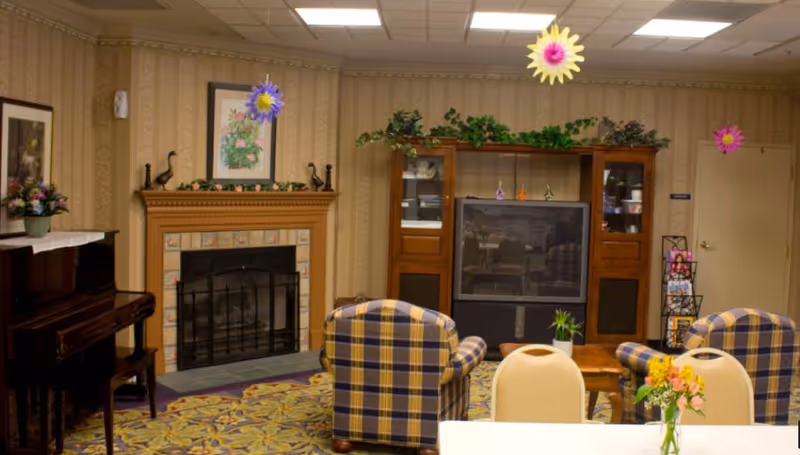 A cozy senior living common area featuring a fireplace with decorative tiles and a mantel adorned with small sculptures and greenery. Two plaid armchairs face a large wooden entertainment center with a television and glass cabinets. A piano with a floral arrangement on top is on the left side. The room has patterned wallpaper, a floral carpet, and hanging colorful paper flowers from the ceiling. A table with chairs and a small vase of flowers is in the foreground.