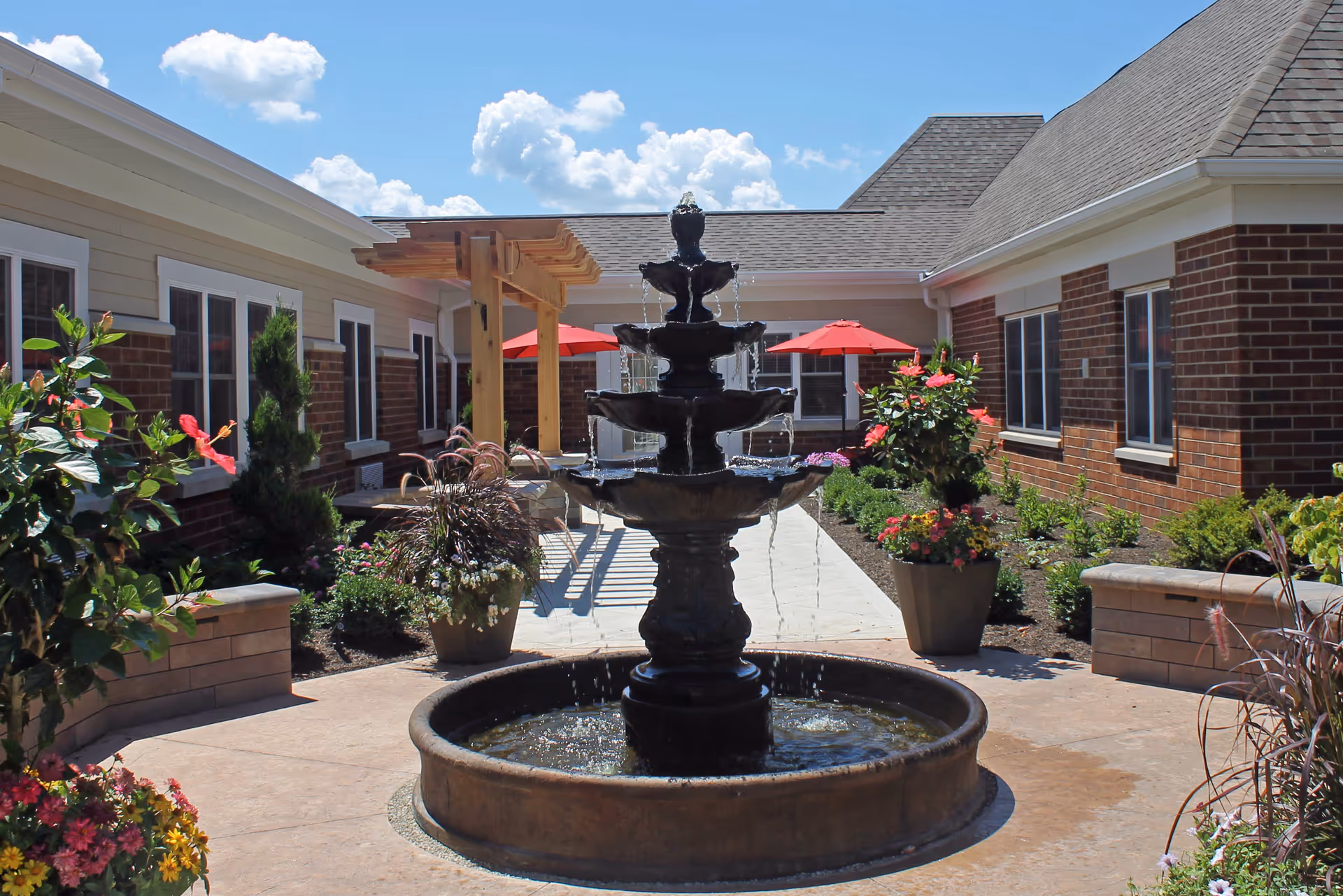 Outdoor courtyard area at Saint Therese at St. Mary of the Woods featuring a multi-tiered black water fountain in the center, surrounded by potted plants and flowers. The courtyard is bordered by brick and siding buildings with windows, and there are red umbrellas providing shade over seating areas in the background under a bright blue sky with scattered clouds.