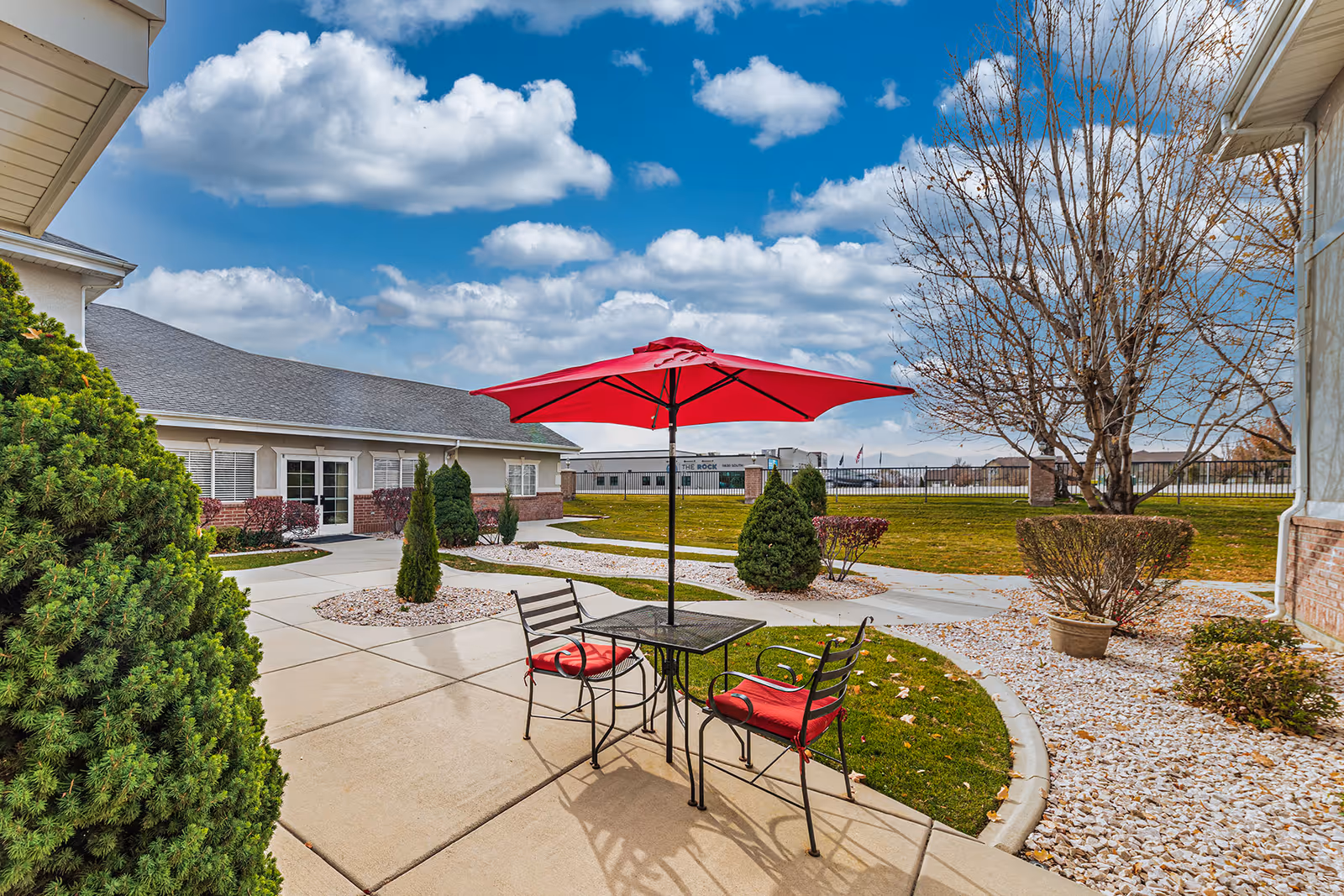 Outdoor patio area with a black metal table and two chairs with red cushions under a red umbrella. The patio is surrounded by landscaped bushes, small trees, and a grassy lawn. A building with white walls and windows is visible in the background under a partly cloudy blue sky.
