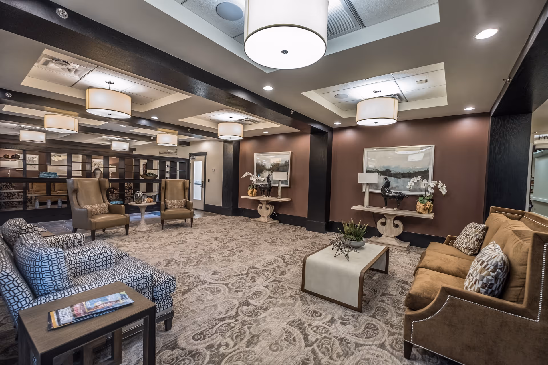 A spacious and elegantly decorated living room area in a senior living facility with patterned carpet, multiple armchairs and sofas in neutral tones, a coffee table with decorative items, wall art, and modern ceiling lights.