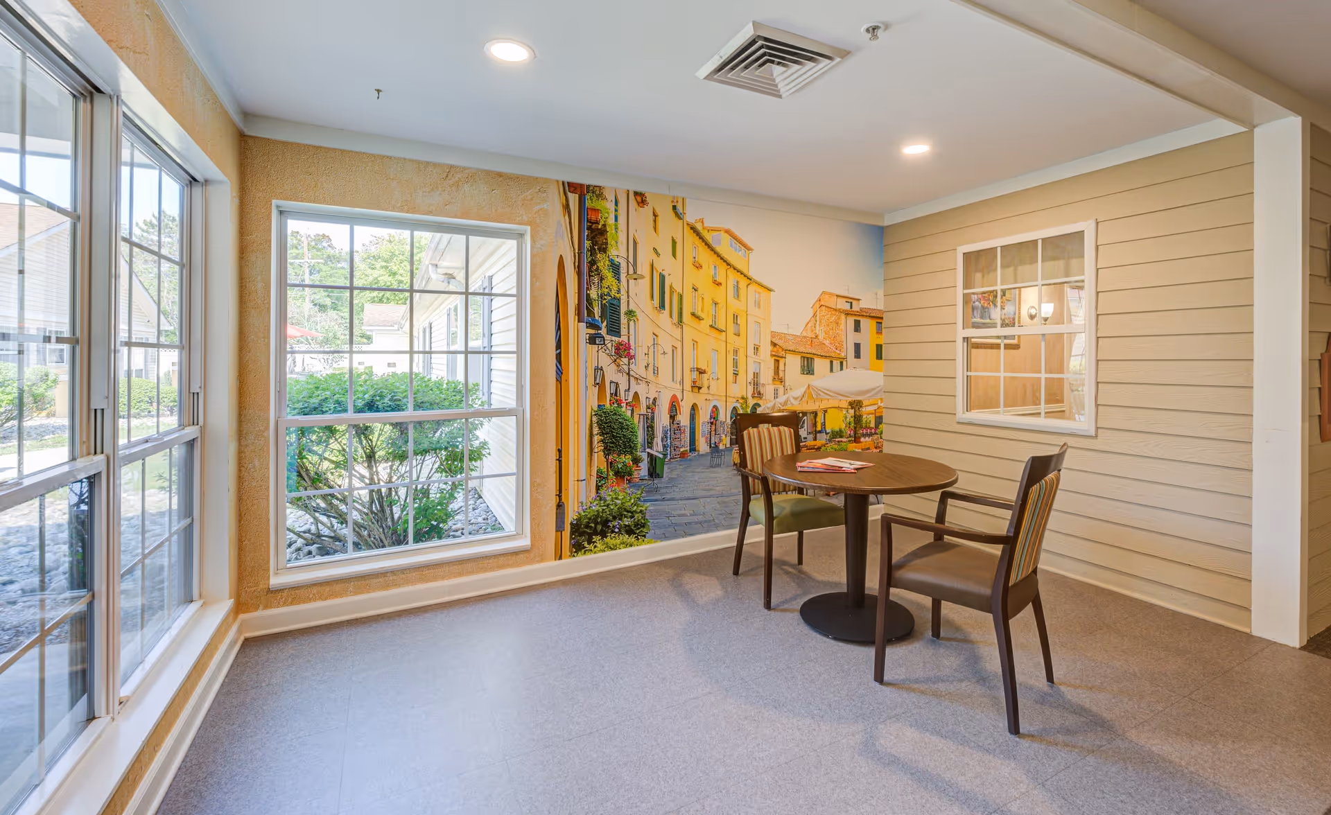 A small seating area in a senior living facility with a round wooden table and two chairs. The room has large windows letting in natural light, a mural of a European street scene on one wall, and beige siding on another wall with a window looking into another room.
