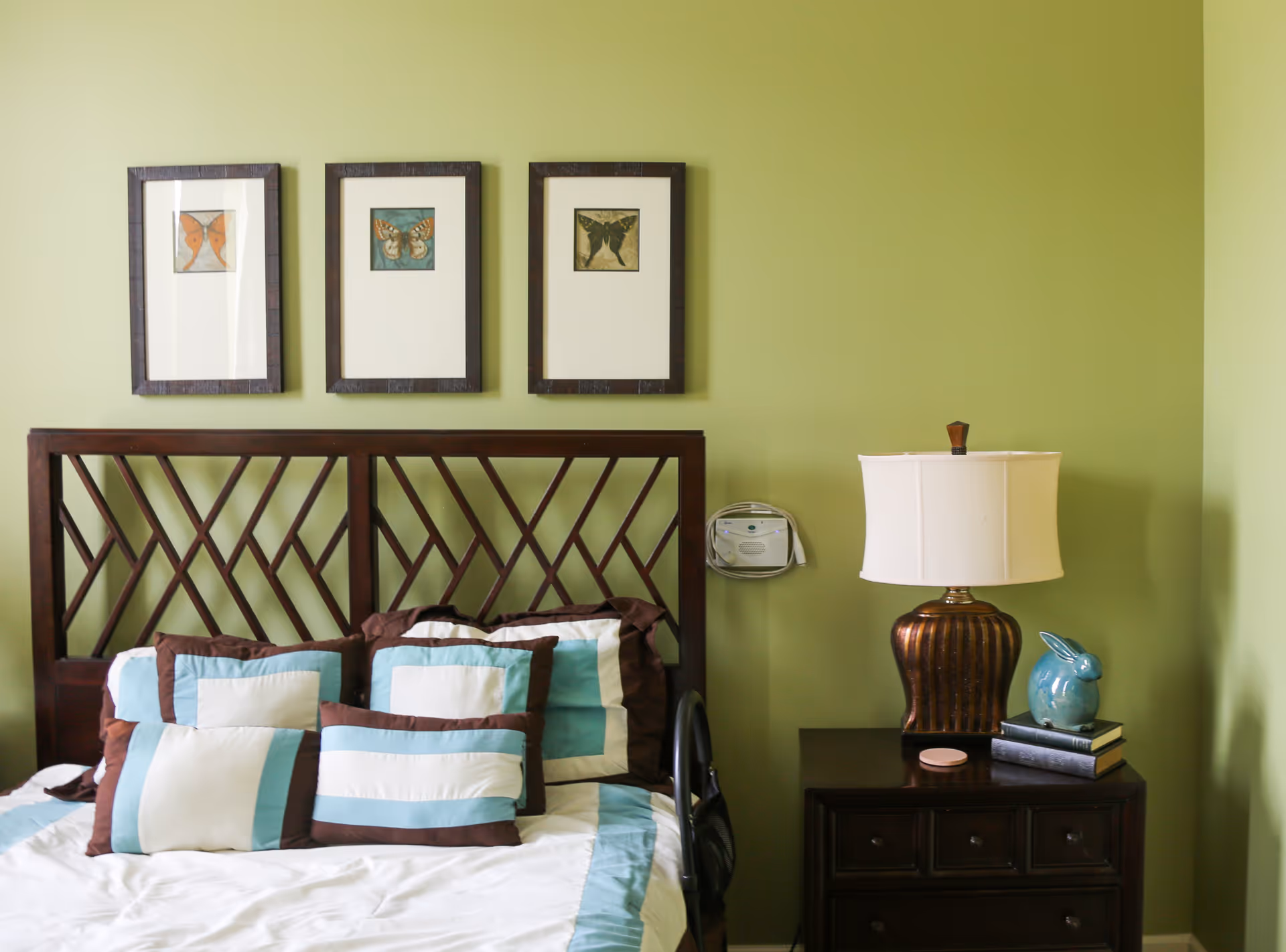 A bedroom with a wooden headboard and a bed made with white, blue, and brown pillows and bedding. Above the headboard are three framed butterfly pictures. To the right of the bed is a dark wooden nightstand with a bronze lamp, a blue ceramic rabbit, and a stack of books. The walls are painted light green.