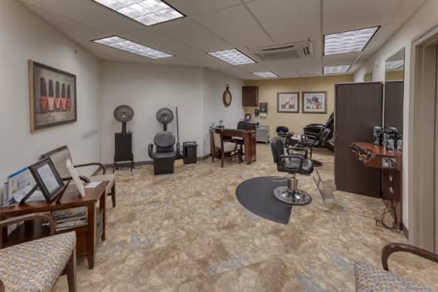 Interior view of a salon area in a senior living facility with salon chairs, hair dryers, a desk, and waiting chairs. The room has beige walls, a tiled floor, framed artwork, and overhead fluorescent lighting.