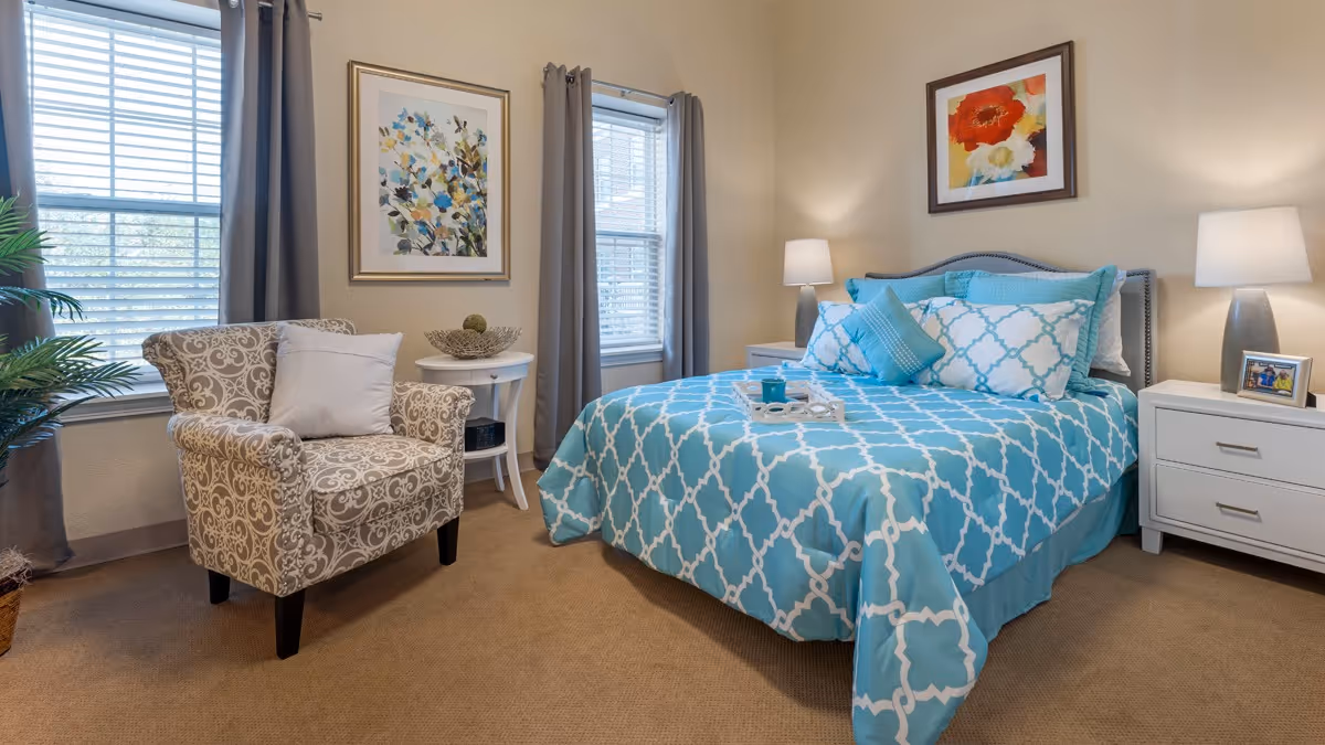 A cozy bedroom with a bed covered in a blue and white patterned comforter and multiple pillows. There are two white nightstands on either side of the bed, each with a lamp. A framed floral painting hangs above the bed. To the left, there is a patterned armchair with a white pillow, a small round white table with decorative items, and two windows with gray curtains allowing natural light to enter the room. A potted plant is partially visible in the corner.