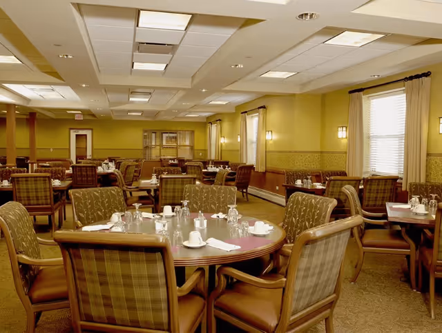 A spacious dining room with multiple round and rectangular tables set with white cups, glasses, and napkins. The room features cushioned chairs with patterned upholstery, yellow walls with decorative trim, and windows with curtains allowing natural light to enter.