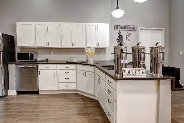 A clean and modern kitchen area with white cabinets, stainless steel refrigerator and dishwasher, a microwave, and a countertop with three large coffee dispensers labeled Decaf, Regular, and Sugar Free. There is also a glass water dispenser with lemon slices on the counter. The walls are painted light gray, and there are coffee-themed decorations on the wall.