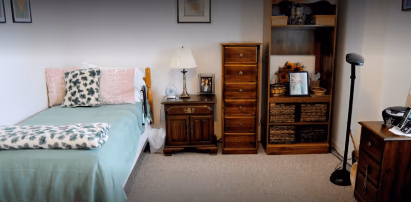 A cozy bedroom with a single bed covered in a green bedspread and floral pillows. Next to the bed is a wooden nightstand with a lamp and framed photo. There is a tall wooden chest of drawers and a wooden bookshelf with baskets and framed pictures. The room has beige carpet and light-colored walls with framed artwork.