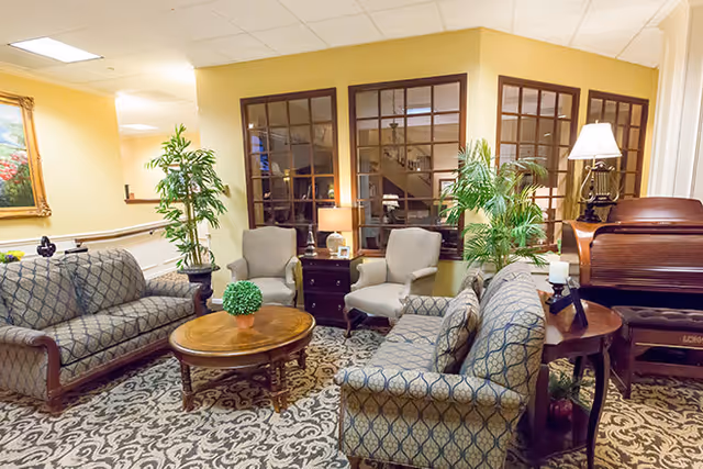 A cozy living room area with two patterned sofas and two armchairs arranged around a round wooden coffee table with a small green plant. The room features a patterned carpet, potted plants, a wooden side table with a lamp, and large windows with wooden frames. There is also a wooden piano with a lamp on top in the corner.