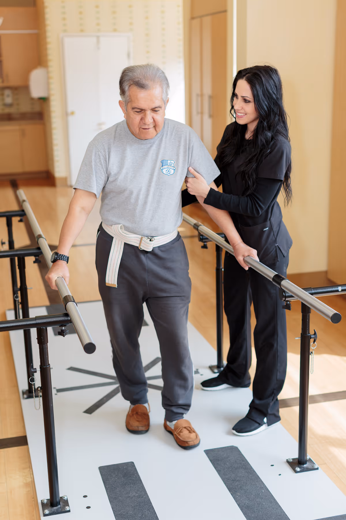 An elderly man wearing a gray t-shirt and dark pants is walking with the support of parallel bars during physical therapy. A female therapist dressed in black scrubs is assisting him by holding his arm and smiling encouragingly. The setting appears to be a rehabilitation facility with wooden floors and light-colored walls.