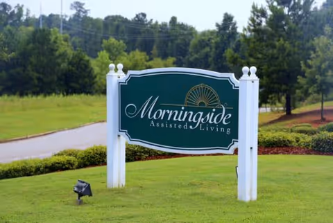 A green and white sign for Morningside Assisted Living standing on a grassy area with trees and a road in the background.