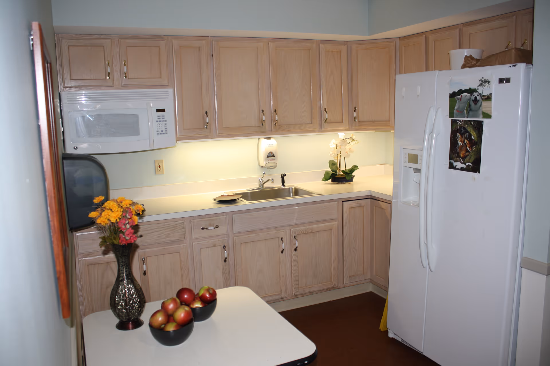 A compact kitchen with light wood cabinets, a white refrigerator and microwave, a sink under-cabinet lighting, and a countertop holding bowls of apples and a vase of flowers.