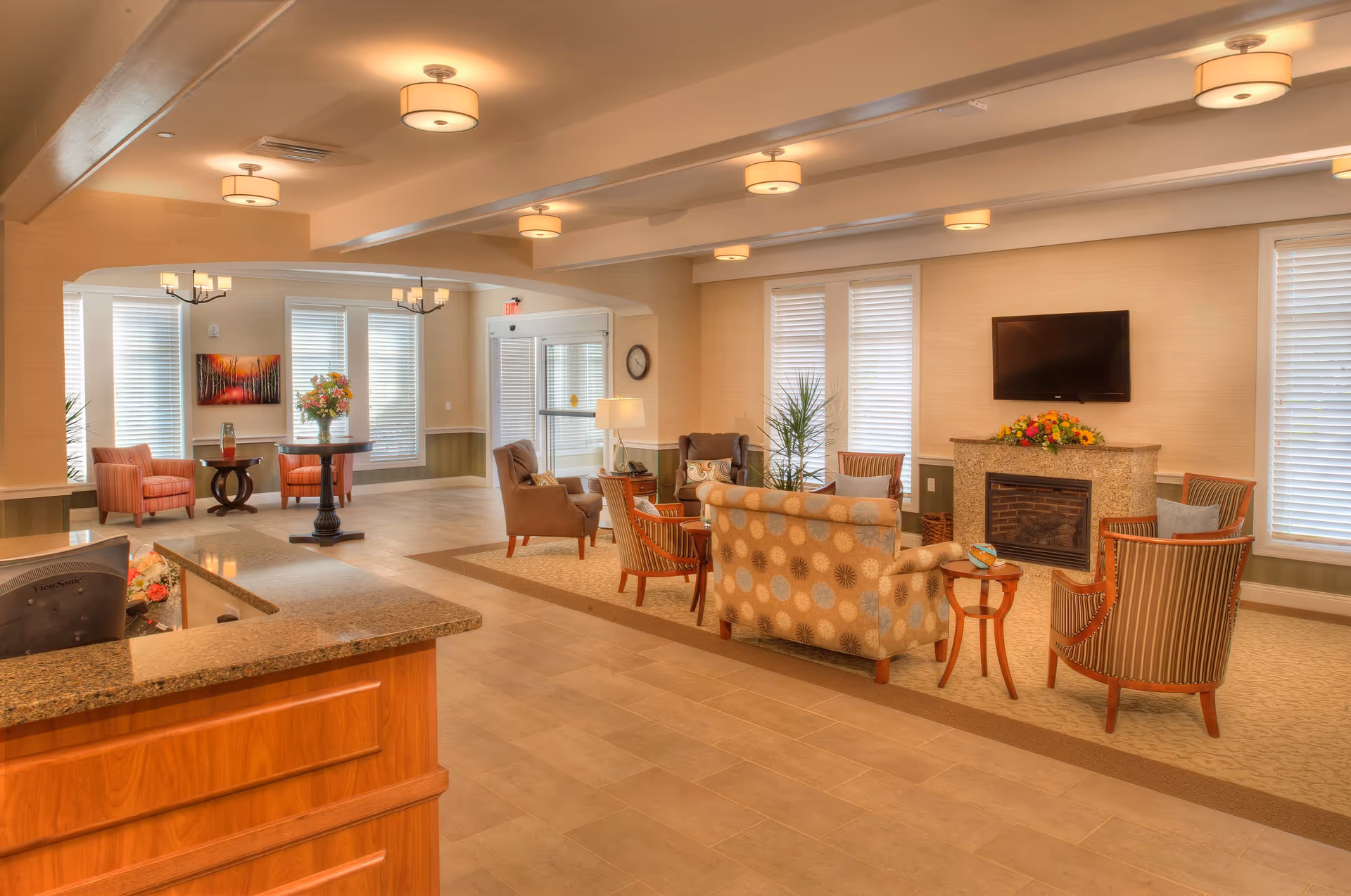 A spacious and well-lit living room area in a senior living facility with comfortable seating including armchairs and a patterned sofa arranged around a fireplace with a flat-screen TV mounted above it. The room features large windows with blinds, a wooden reception desk with a granite countertop, decorative plants, and warm lighting fixtures on the ceiling.