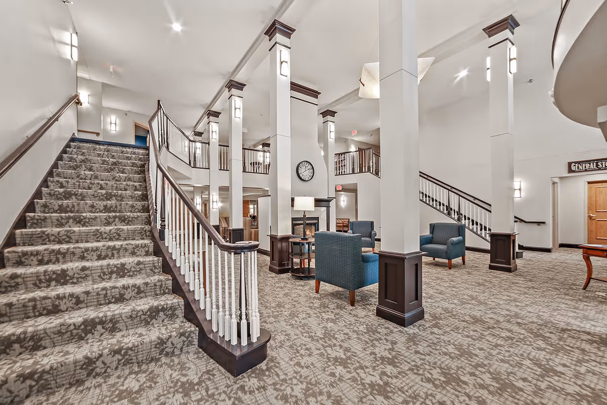 Spacious interior of a retirement community featuring a carpeted staircase with patterned carpeting, white pillars with dark wood accents, blue upholstered chairs arranged in seating areas, a wall clock, and multiple light fixtures illuminating the space.