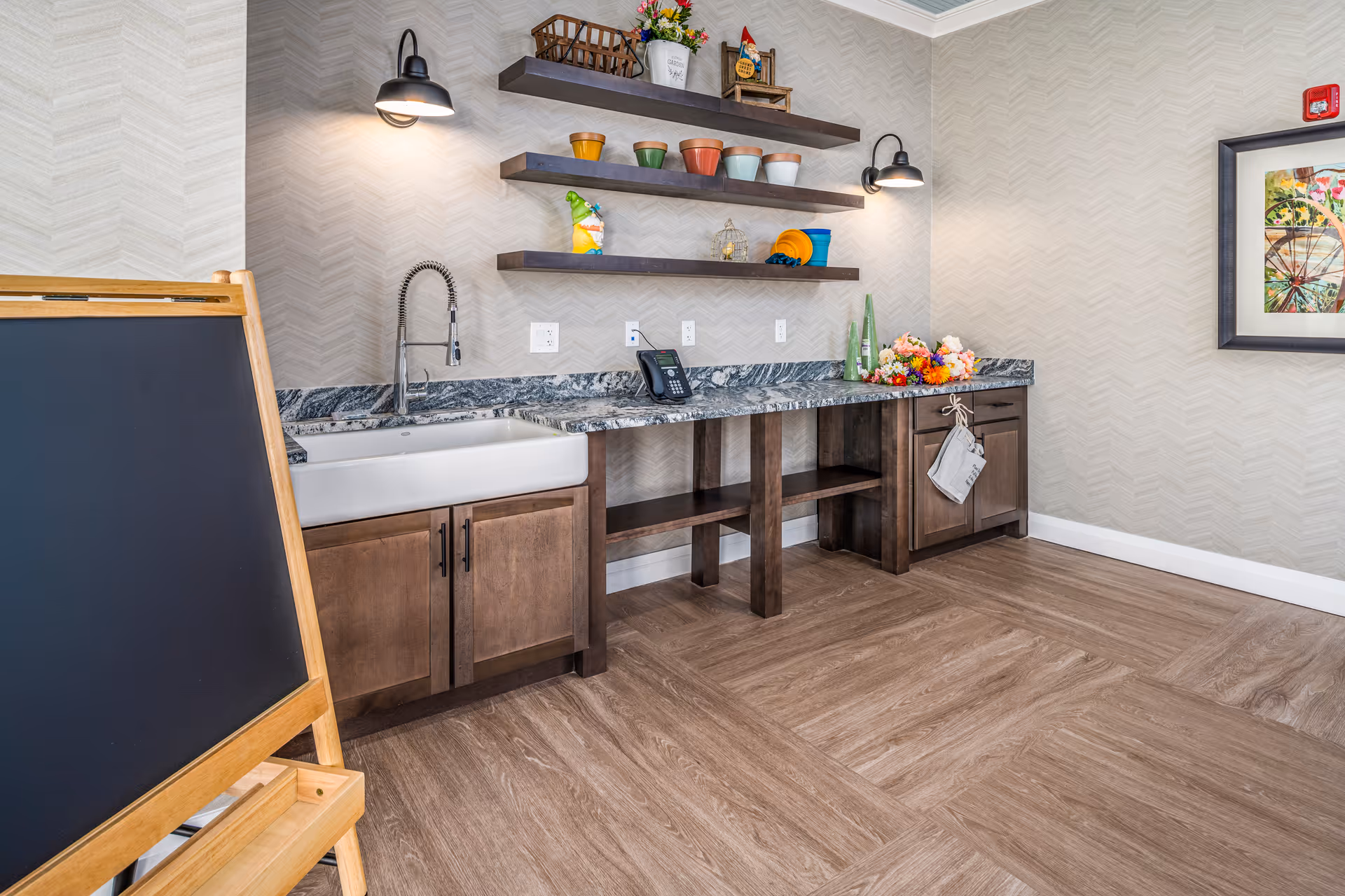 A room with a long countertop featuring a farmhouse-style sink, dark wood cabinets, and three floating shelves holding colorful pots and decorative items. Two wall-mounted lights illuminate the area. A blackboard on a wooden easel is positioned on the left side, and a framed artwork hangs on the right wall. The floor has a wood-like finish, and the walls have a subtle chevron pattern wallpaper.