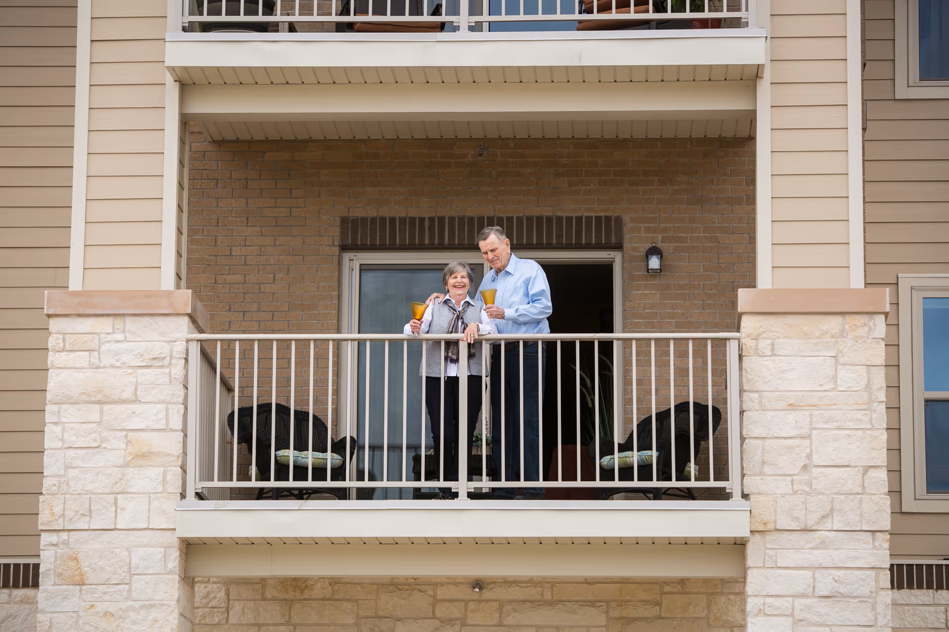 An elderly couple standing on a balcony of a residential building, holding drinks and smiling. The balcony has two chairs with cushions and is surrounded by a metal railing. The building exterior features brick and stone walls.