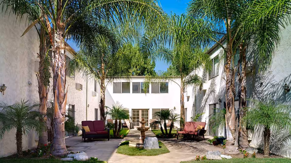 Outdoor courtyard area at Atria Covina featuring tall palm trees, a central stone fountain, green grass patches, and cushioned seating benches against white building walls under a clear blue sky.