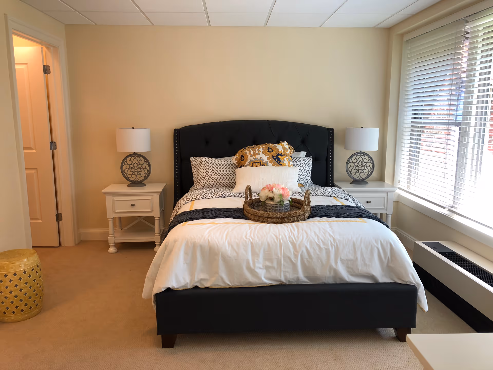 A neatly made bed with a dark upholstered headboard and multiple pillows, flanked by two white nightstands each with a decorative lamp. A woven tray with flowers sits on the bed. To the right, there is a large window with white blinds, and to the left, a door is partially open revealing a lit room.