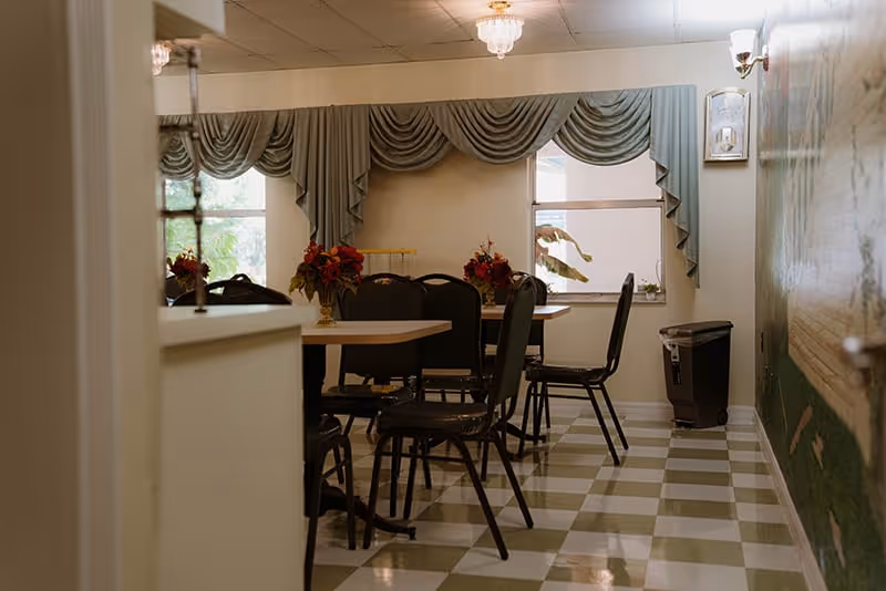 Small dining room with tables and chairs, floral centerpieces, draped valances over windows, and a checkered tile floor.