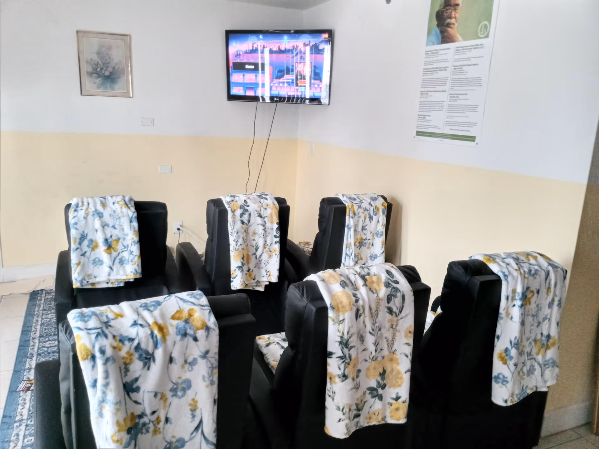 A small common area with six black leather chairs arranged in two rows facing a wall-mounted TV. Each chair has a white blanket with yellow and blue floral patterns draped over the back. The walls are painted white with a beige lower half, and there are two framed pictures hanging on the walls.