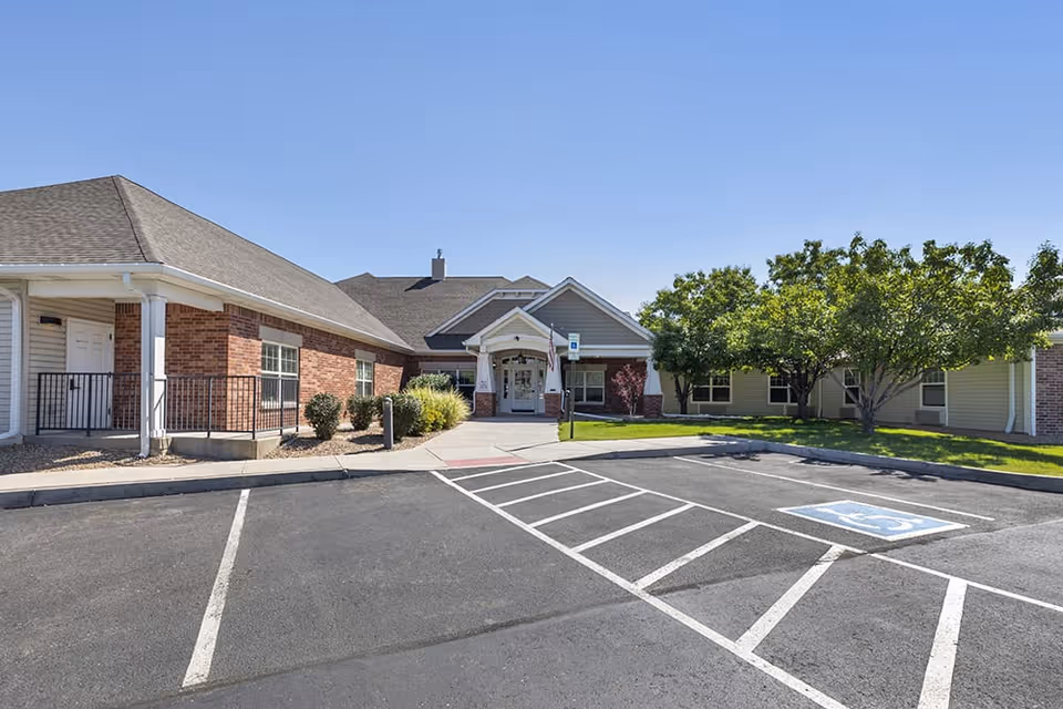 Exterior view of Brookdale Arvada facility showing a single-story building with a combination of brick and siding. There is a paved parking lot with marked handicap parking spaces in front, and a sidewalk leading to the main entrance. Trees and shrubs are visible around the building under a clear blue sky.