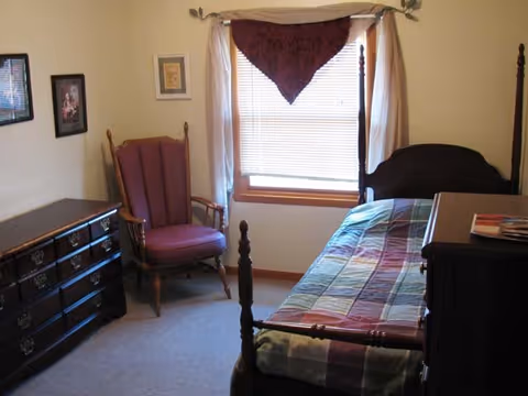 A small bedroom with a wooden four-poster bed covered with a multicolored quilt, a wooden dresser with multiple drawers, a wooden armchair with red upholstery, and a window with blinds and a red valance.