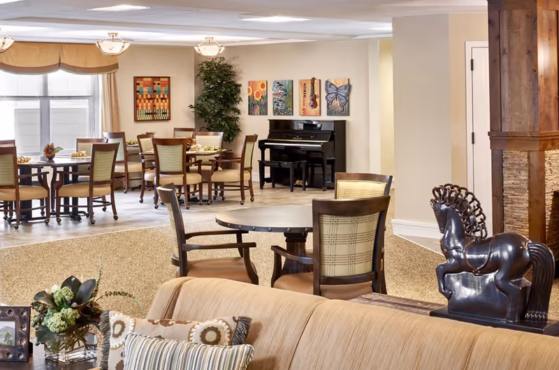 A spacious and well-lit common area in an assisted living facility featuring multiple round tables with chairs, a black upright piano against the wall, decorative paintings above the piano, a large potted plant, and a comfortable beige sofa with patterned pillows in the foreground. There is also a decorative horse sculpture on a wooden surface near a stone and wood column.