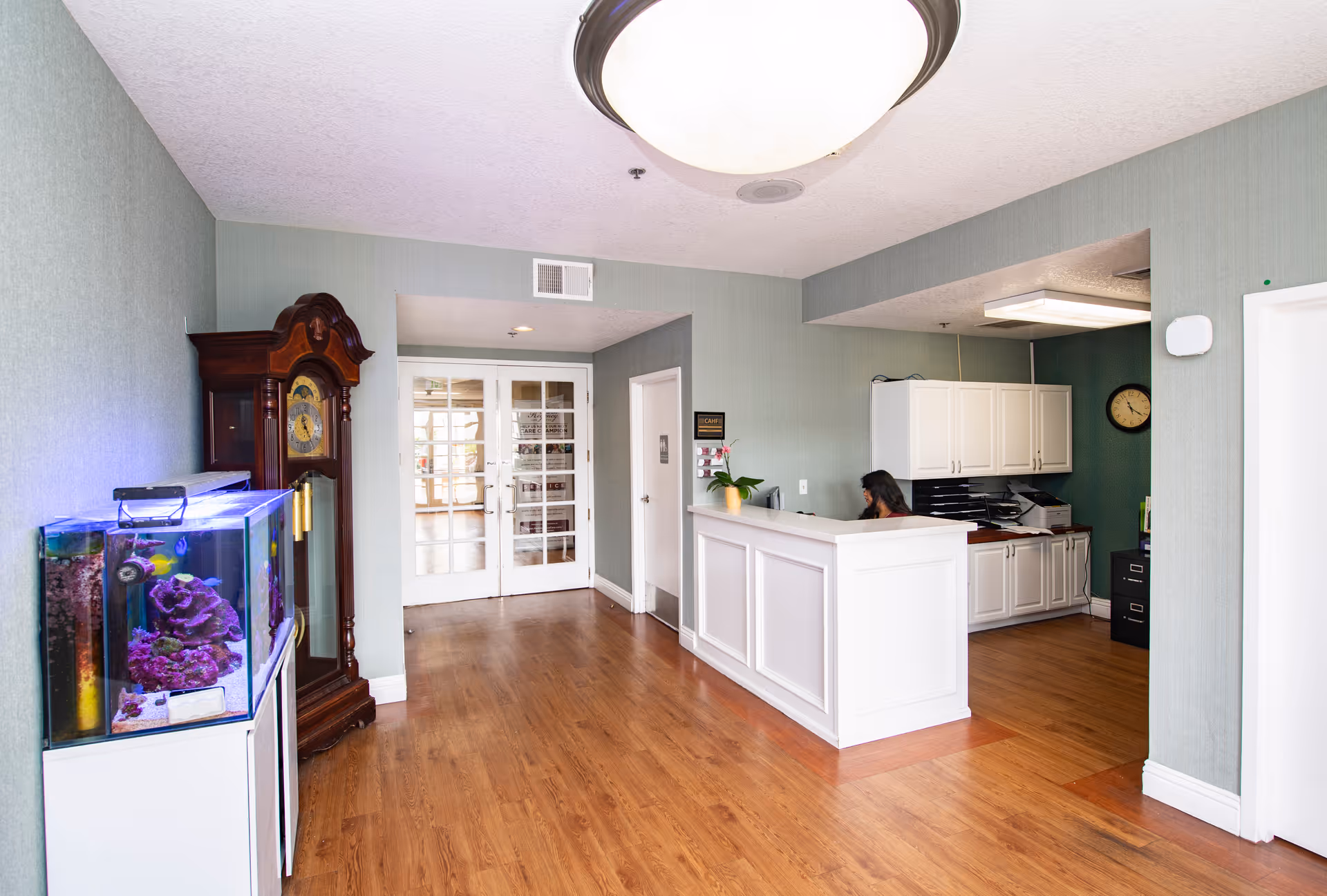 Reception area of a senior living facility with a white check-in desk, a fish tank, a grandfather clock, and a receptionist at the counter.