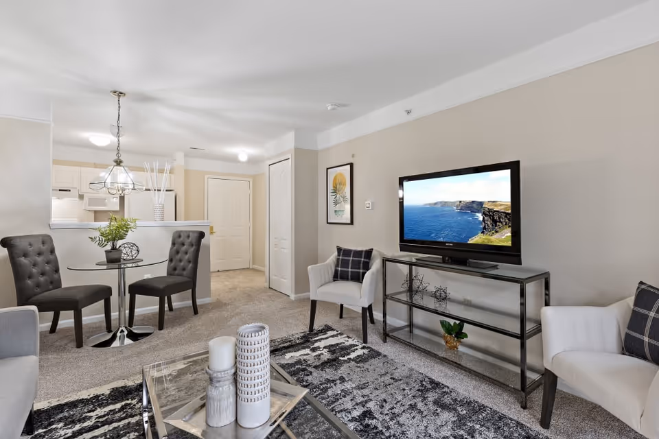 A bright and modern living room area with a flat-screen TV on a glass and metal stand, two white armchairs with plaid pillows, a glass coffee table with decorative candles, and a small round glass dining table with two dark upholstered chairs. The room has beige walls, carpeted floors, and a view into a kitchen area with white cabinets and appliances.