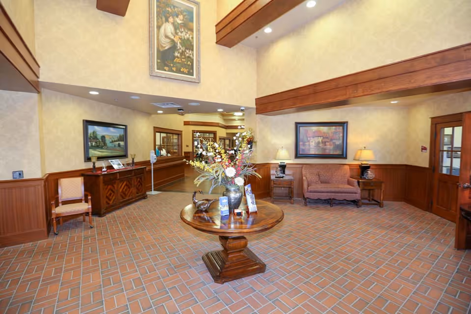 A spacious and well-lit nursing home lobby with a round wooden table in the center holding a large floral arrangement and brochures. The room features brick flooring, wood paneling on the walls, framed paintings, a cushioned loveseat with side tables and lamps, and a wooden cabinet with a chair beside it. The ceiling has wooden beams and recessed lighting.