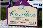 Outdoor sign for Carillon Nursing and Rehabilitation Center with the address number 830, set on a grassy area with a white rock border and a parked white vehicle in the background.
