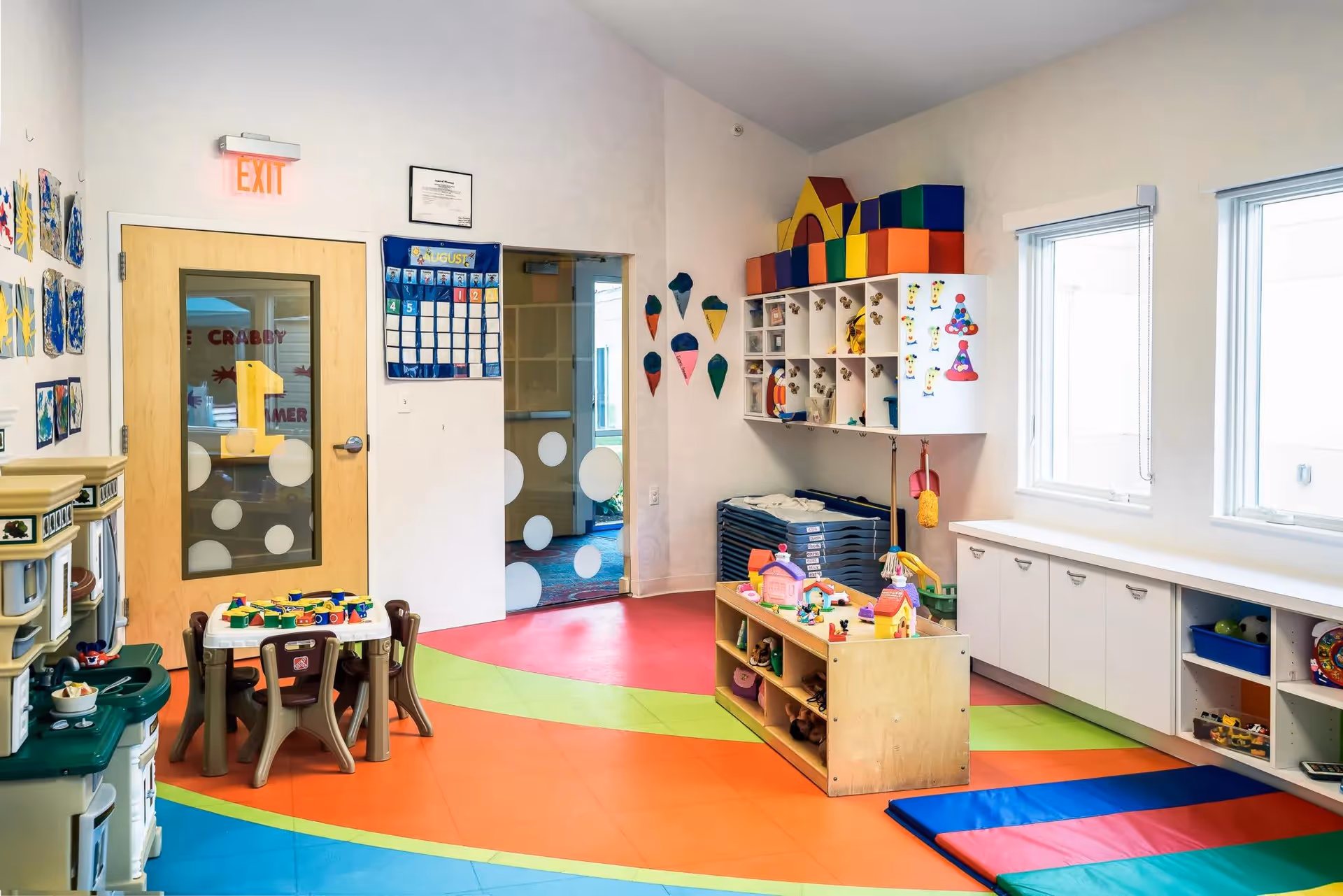 A brightly colored playroom with a multicolored floor, small tables and chairs, toy kitchen set, shelves with toys and stuffed animals, and wall decorations including a calendar and ice cream cone cutouts. Two windows let in natural light.