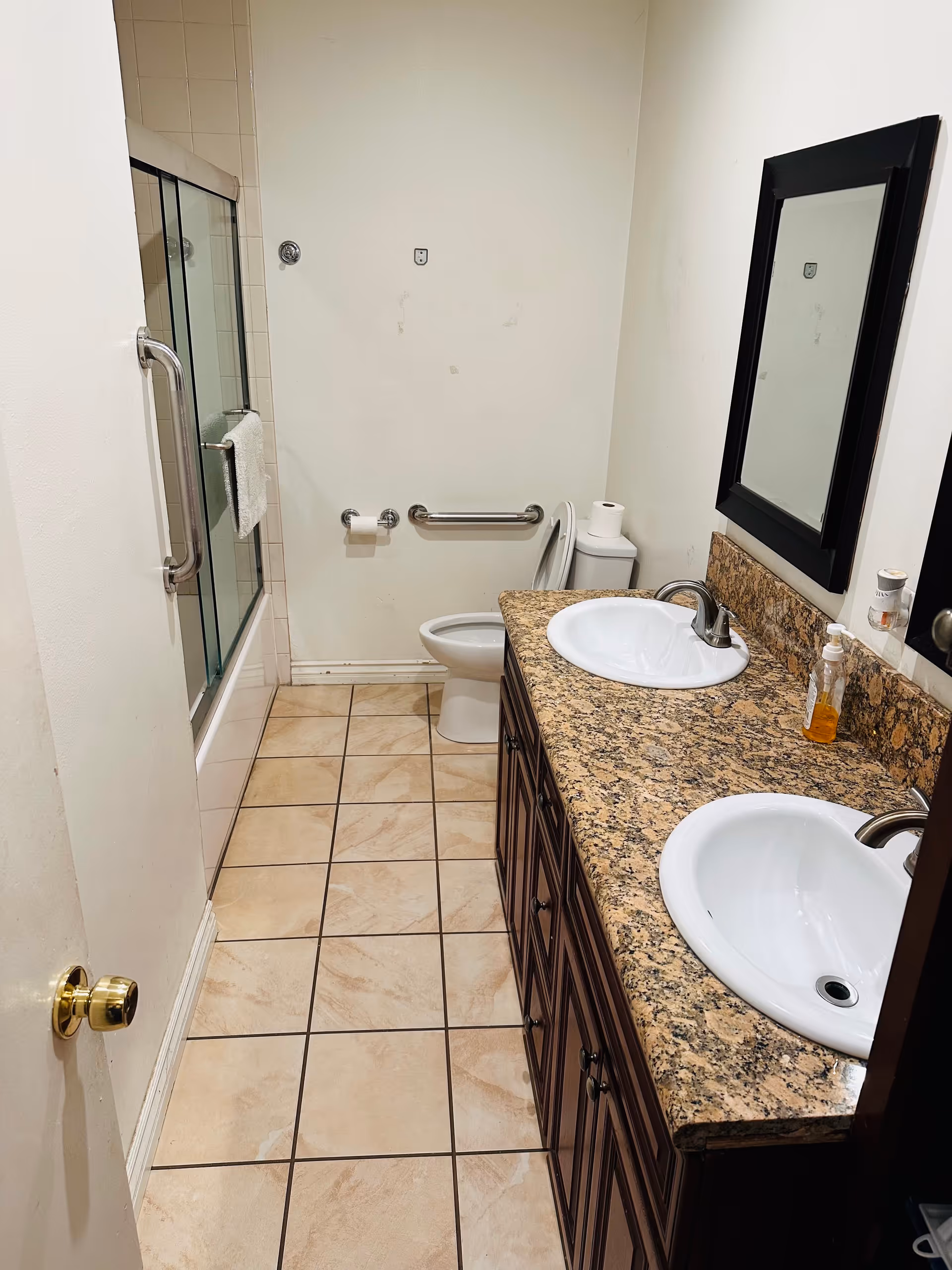 Bathroom with double sinks on a granite vanity, toilet with grab bar, and a bathtub with sliding glass doors.