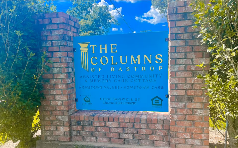 Blue sign reading The Columns of Bastrop between brick pillars at the facility entrance.
