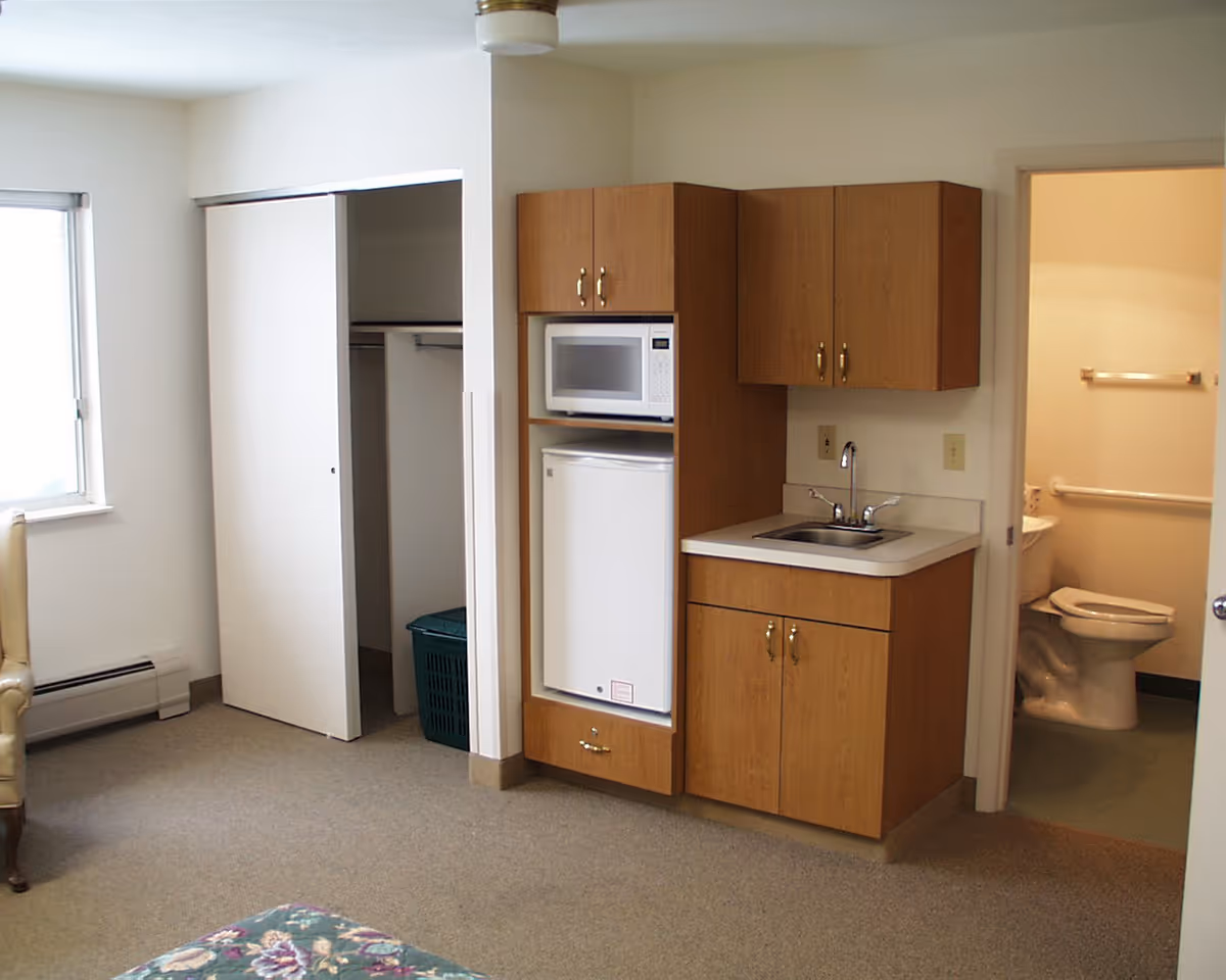Interior view of a room in an assisted living facility showing a small kitchenette with a microwave, mini refrigerator, and sink. To the right, there is a doorway leading to a bathroom with a toilet and grab bars. On the left side, there is an open closet with sliding doors and a window letting in natural light.