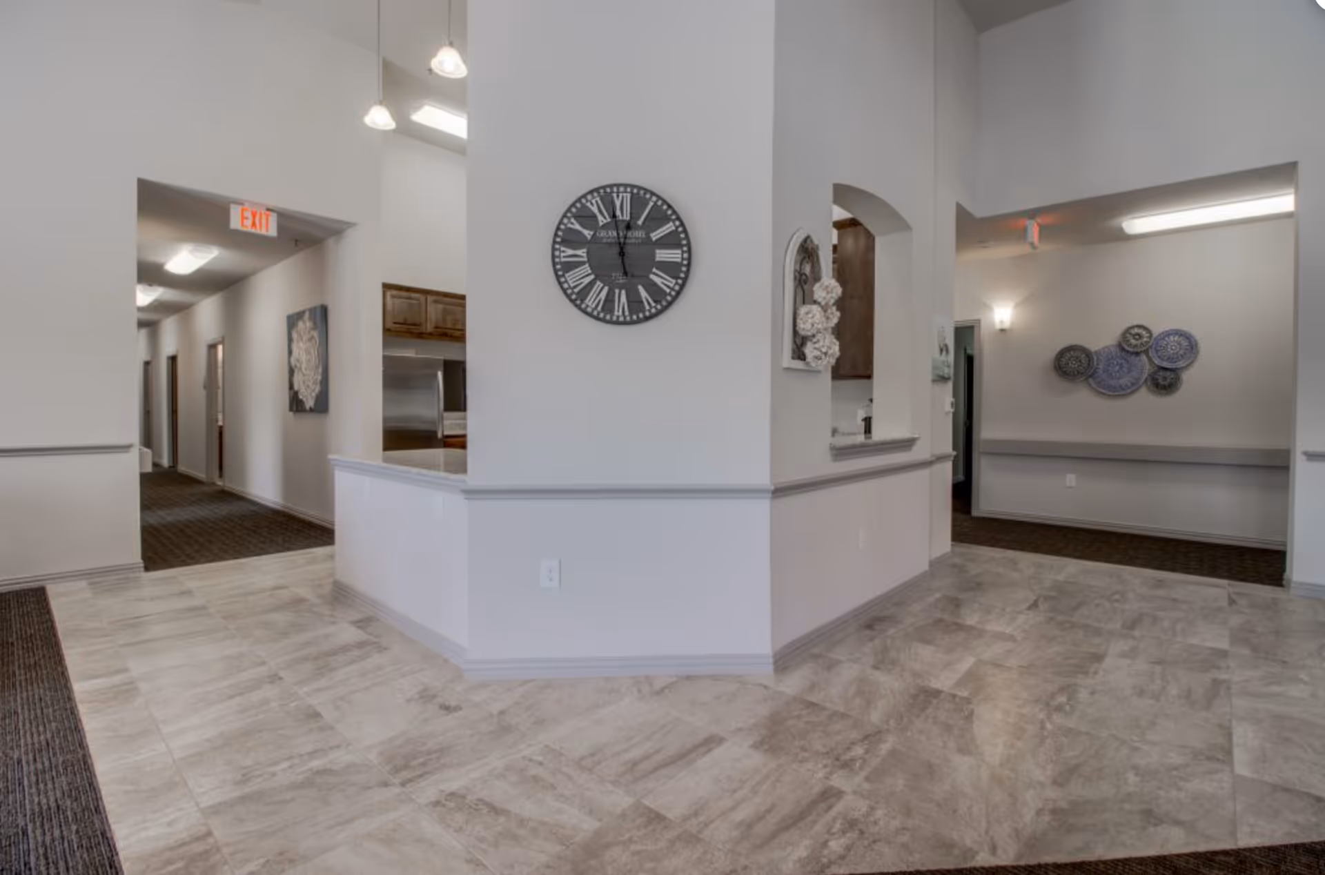 Bright assisted living facility reception area with tiled floor, a central partial wall featuring a large clock, hallways, and decorative wall art.