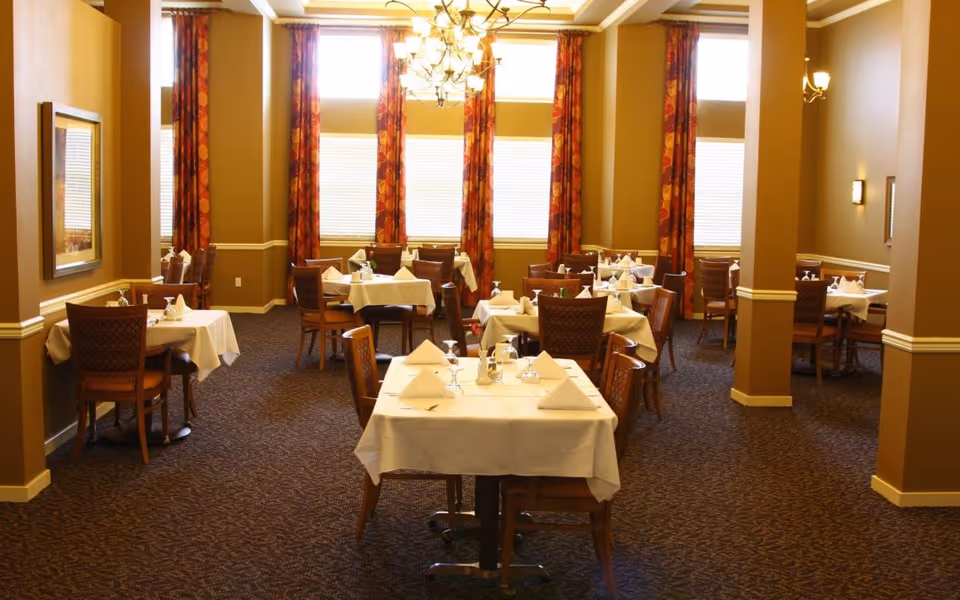 Formal dining room with multiple tables set with white tablecloths, chairs, and folded napkins under chandeliers and tall windows.