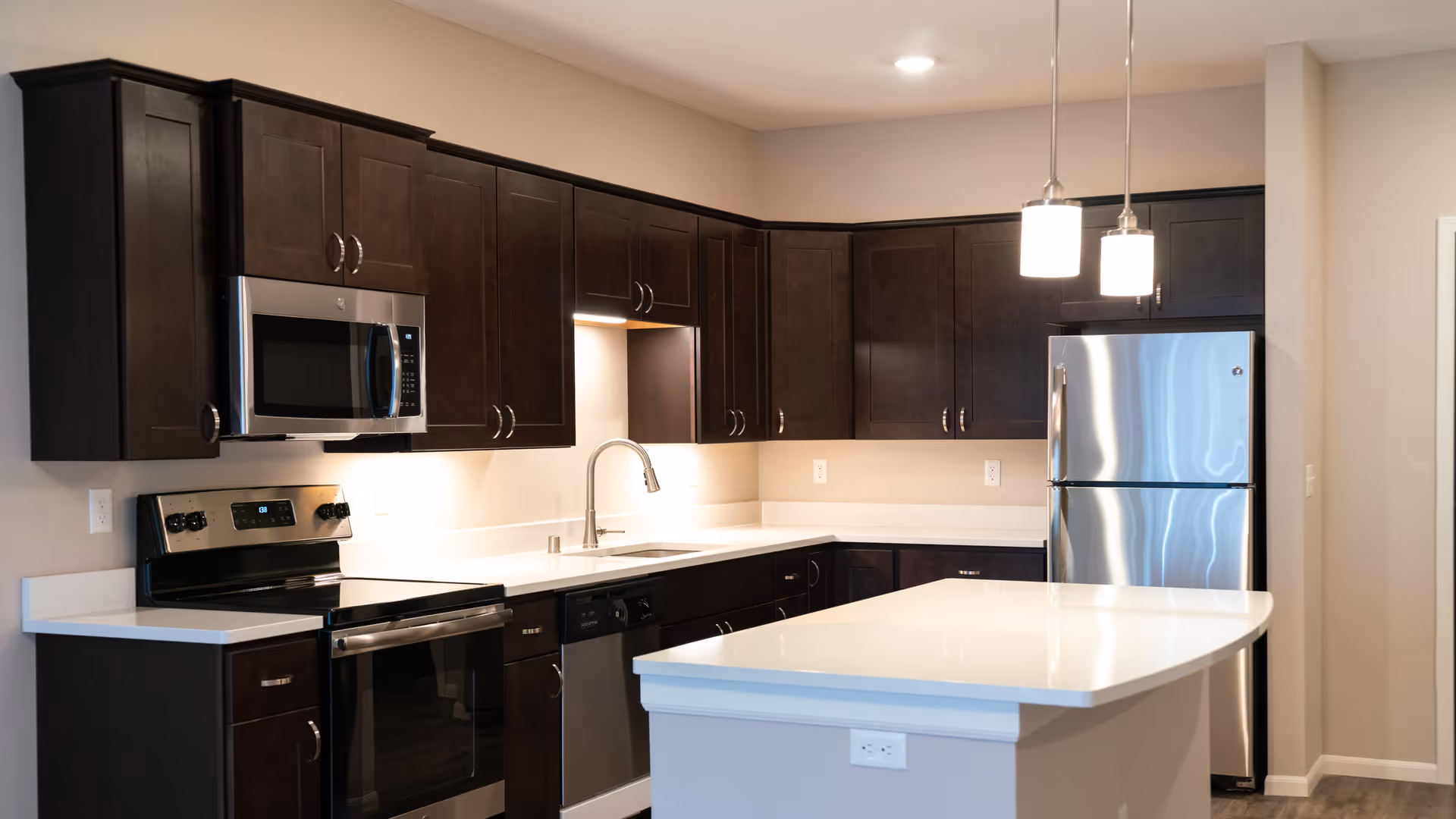 Modern kitchen with dark wooden cabinets, stainless steel appliances including a microwave, oven, dishwasher, and refrigerator, white countertops, a sink with a curved faucet, and two pendant lights hanging above a kitchen island.