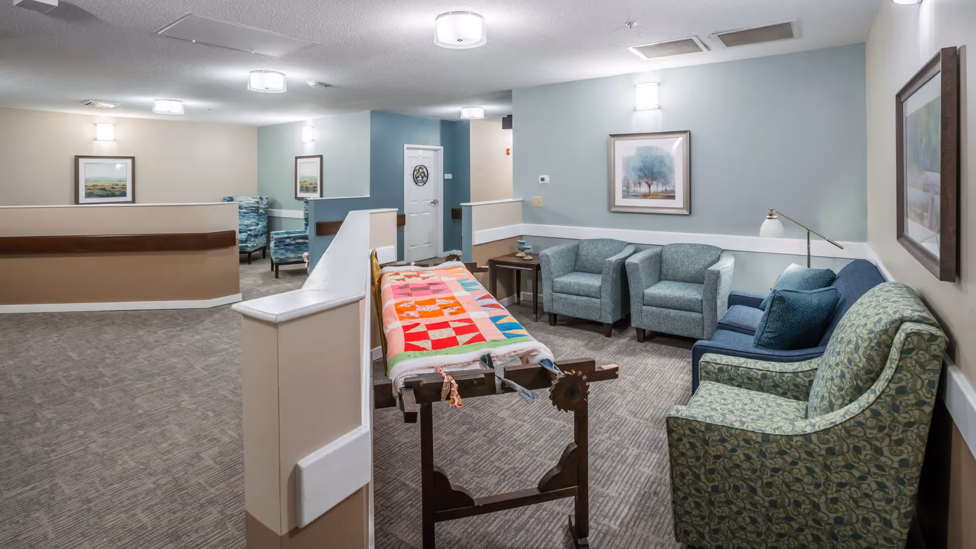A cozy seating area in a senior living facility with several upholstered armchairs and a couch arranged around a small wooden table. A colorful quilt is draped over a wooden quilt rack in the foreground. The walls are painted in soft beige and blue tones, decorated with framed artwork and wall-mounted lights. The carpeted floor and partial room dividers create a warm and inviting atmosphere.